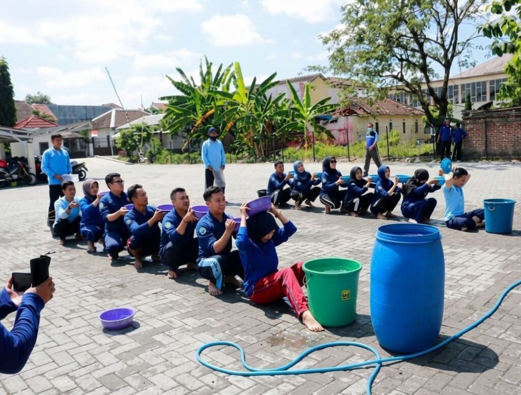 Peserta lomba estafet air memindahkan air menggunakan gelas plastik sebagai permainan kelompok yang melatih kerja sama dan koordinasi.